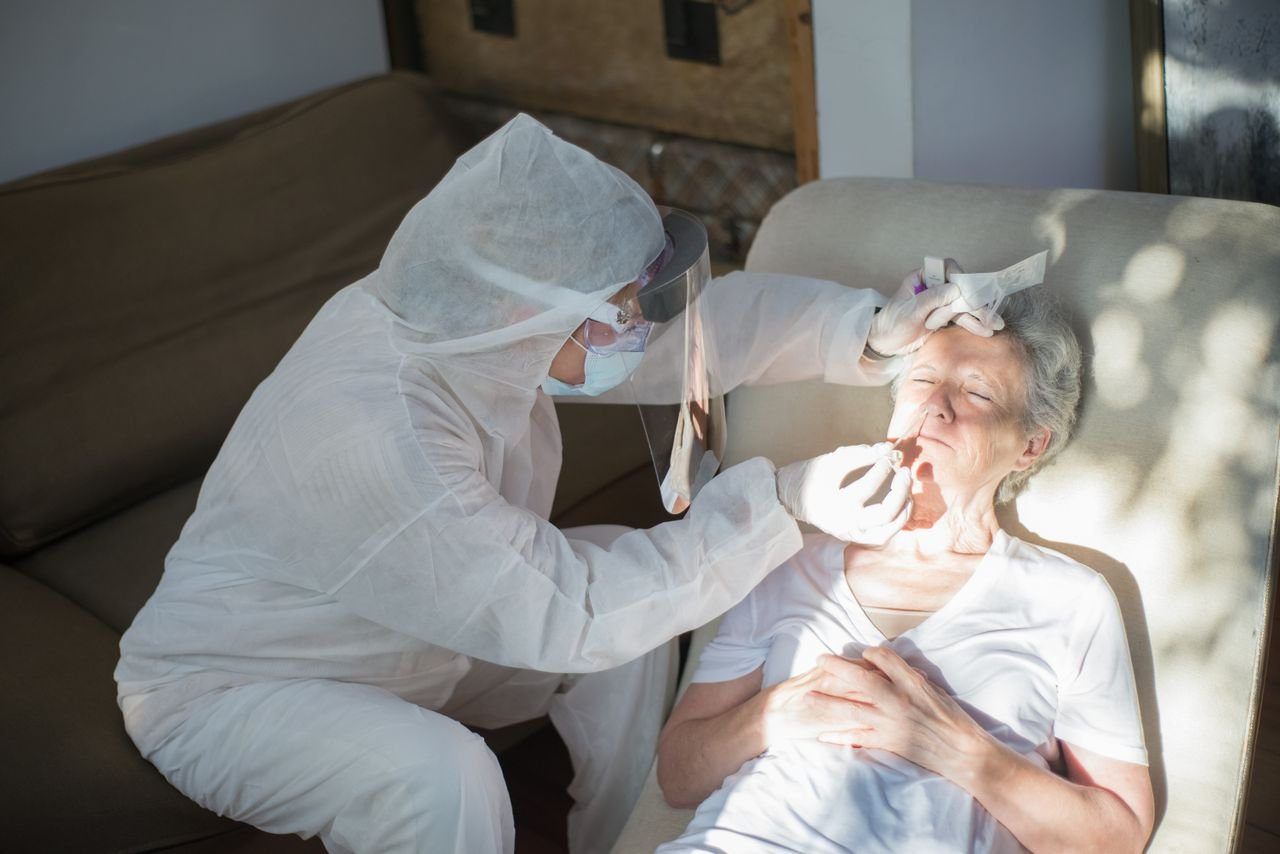 about-02 A healthcare professional in protective gear performs a COVID-19 test on a senior woman at home.